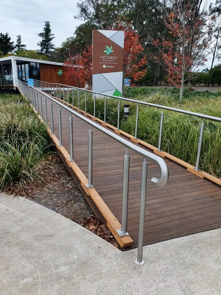 Wooden accessibility ramp with metal handrails leading to a building surrounded by greenery and autumn-colored trees, with a sign that reads The Gables Box Hill.