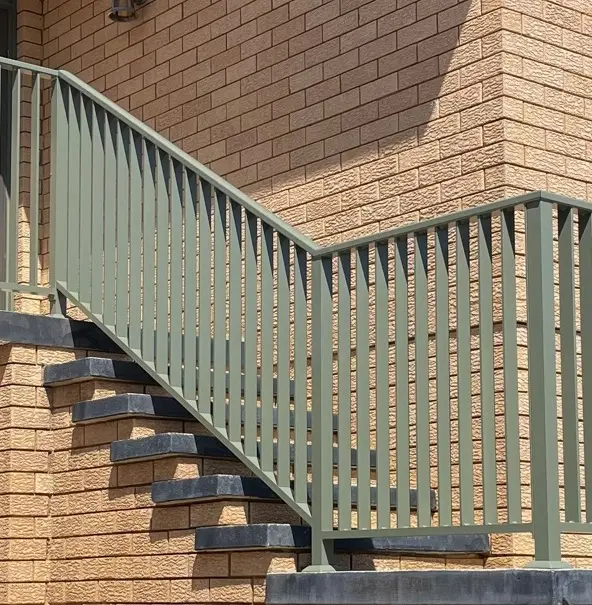 Outdoor staircase with dark stone steps and a green metal railing attached to a textured beige brick wall.