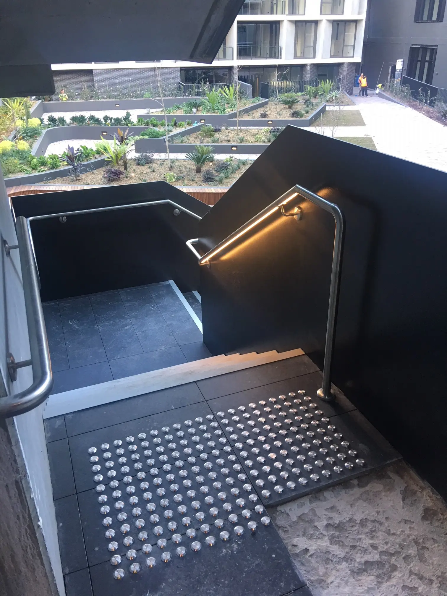 Staircase with tactile paving, metal handrails, and a lit handrail on a modern building, overlooking landscaped garden beds.