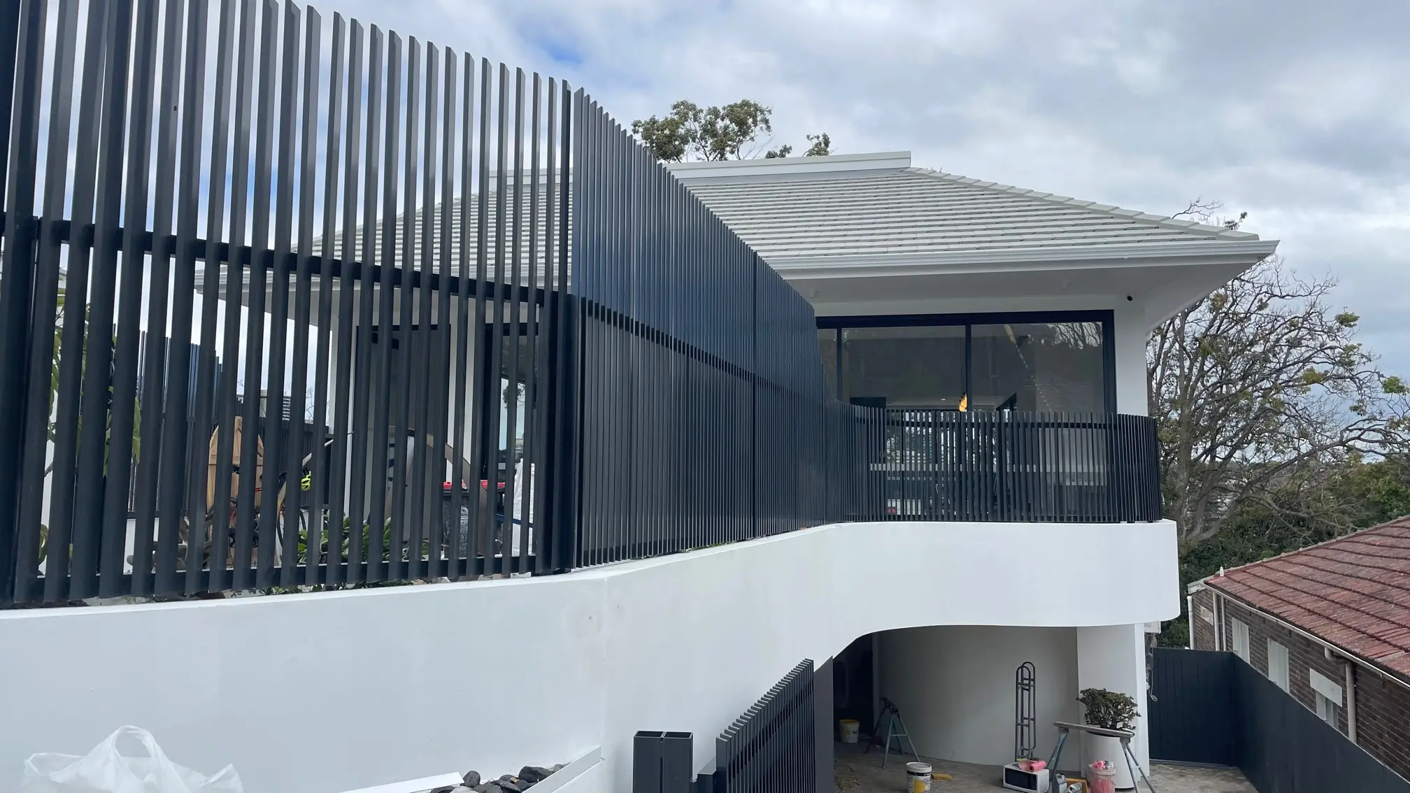 Modern two-story house with a grey tiled roof, black vertical metal fence, large window, and a small balcony on a cloudy day.