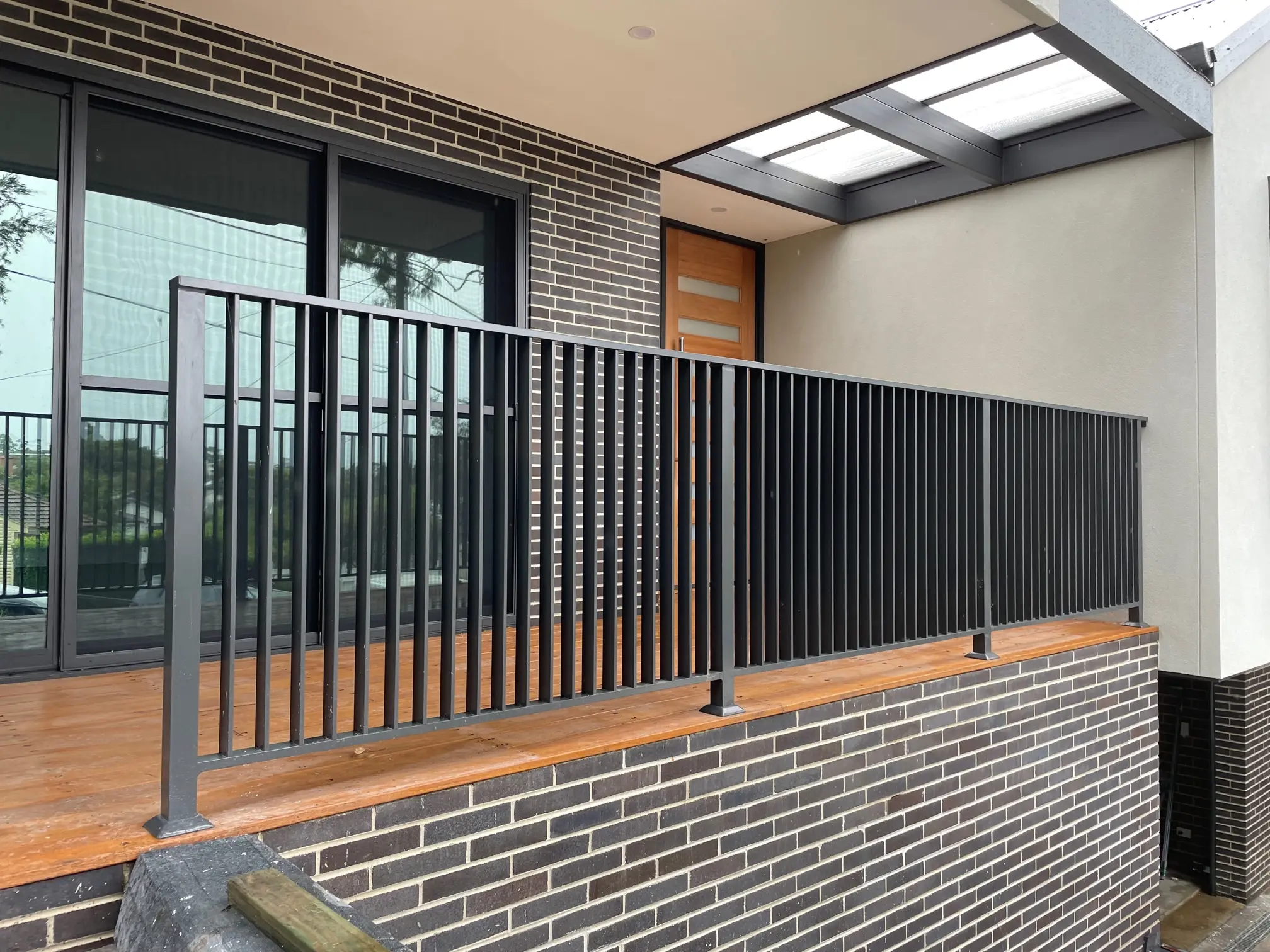 Modern house balcony with black vertical metal railing, wooden floor, dark brick wall, and wooden door under a transparent roof.