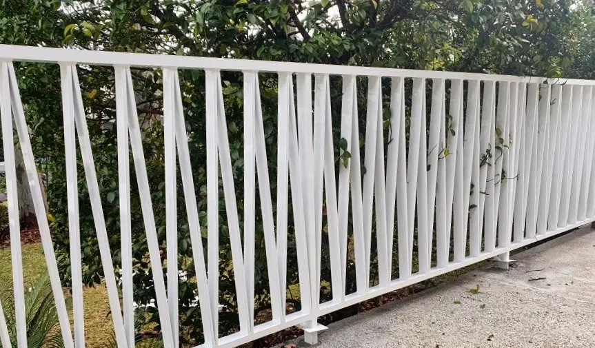White metal railing with vertical bars forming a zigzag pattern in front of green leafy bushes.