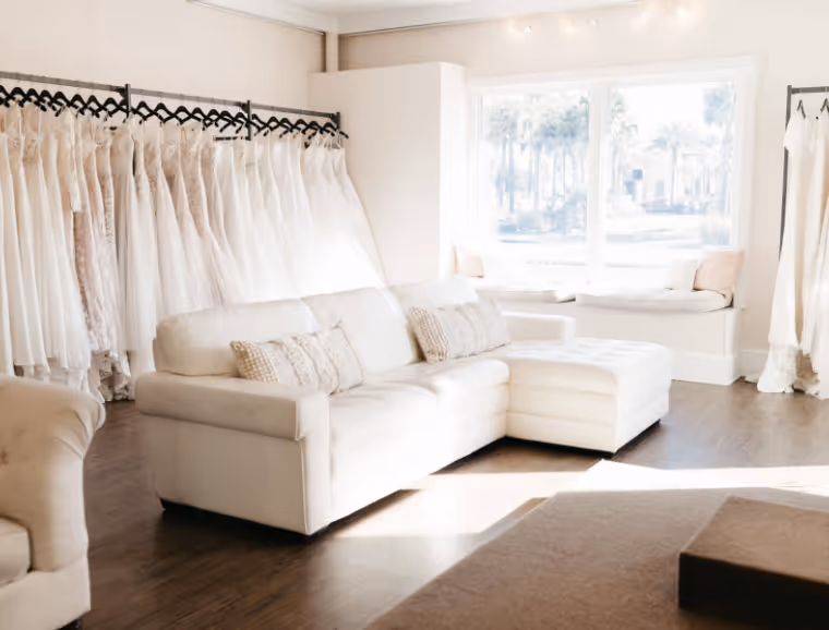 Bright bridal shop interior with a white sectional sofa, wooden floor, racks of white wedding dresses, and a large window seat with cushions.
