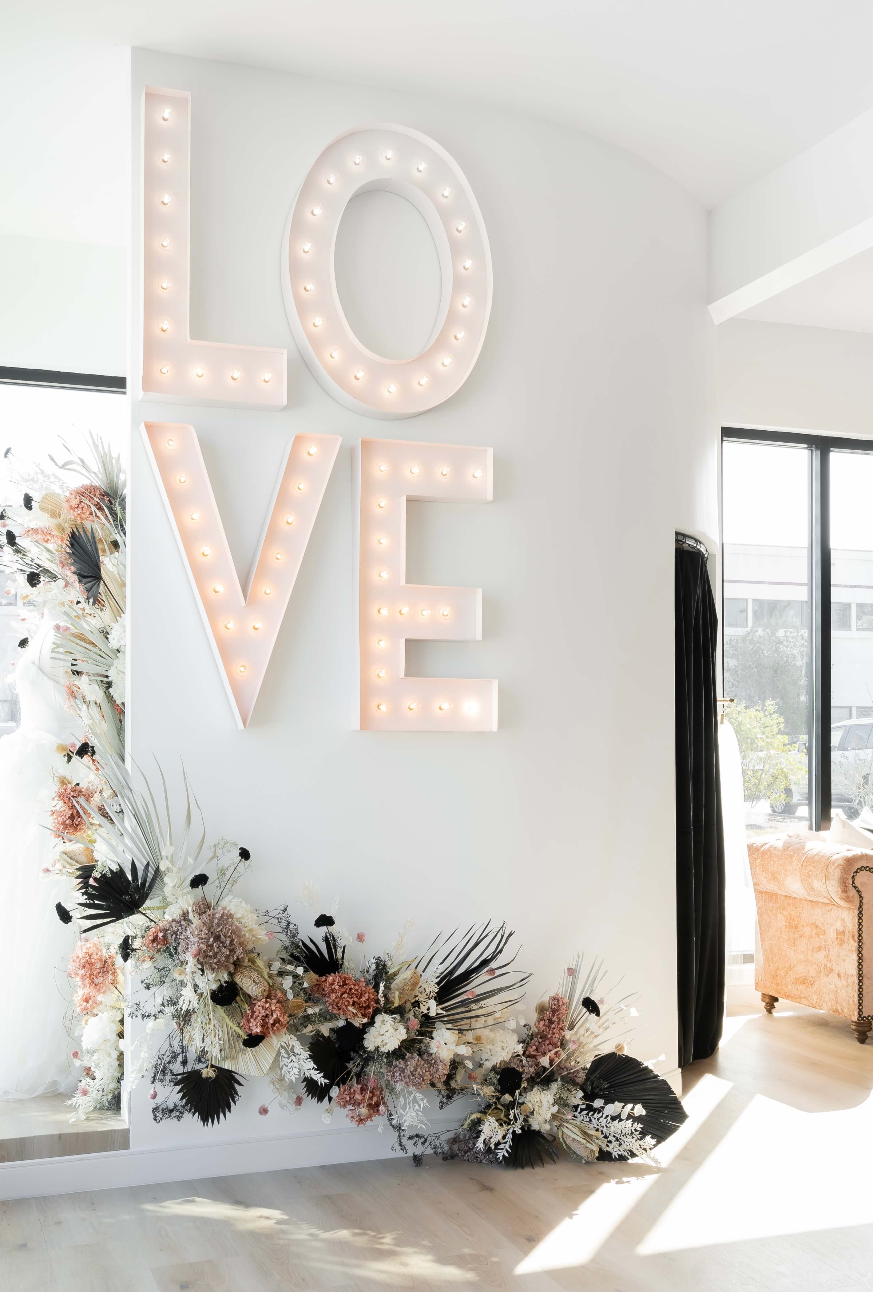 Large illuminated letters spelling LOVE on a white wall with a floral arrangement at the base in a bright room.