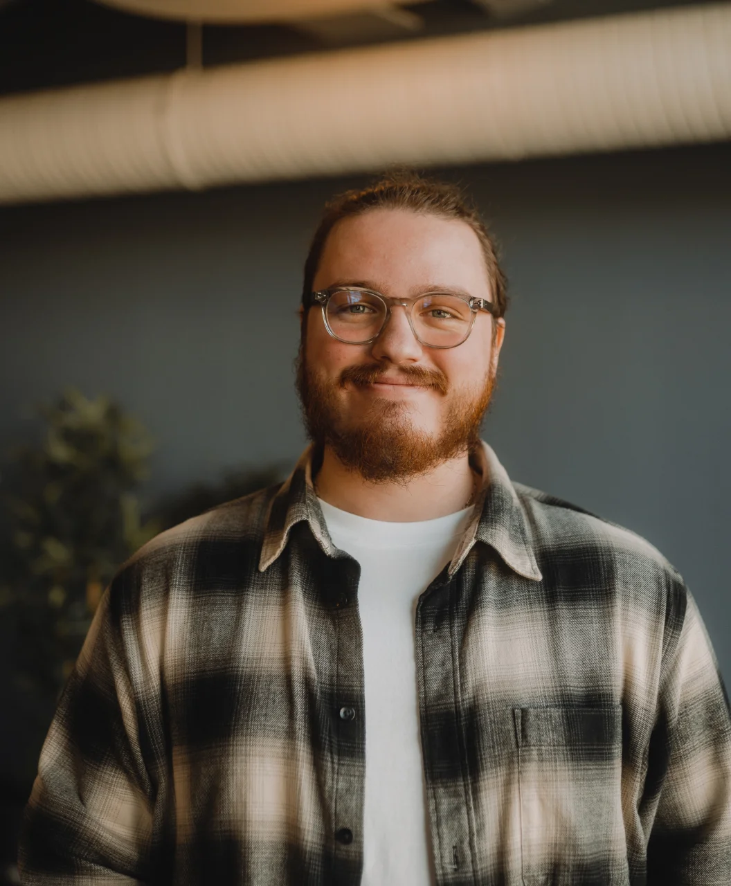 A person with glasses and a beard smiles while wearing a plaid shirt.