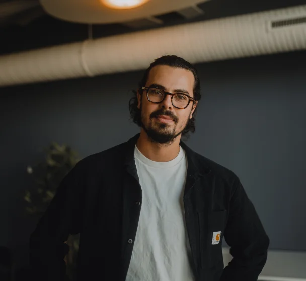 A person wearing glasses and a pink sweater smiles slightly, standing indoors.
