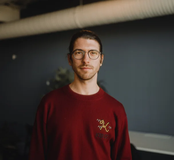 A man with glasses and a beard stands indoors, wearing a black jacket and white shirt.