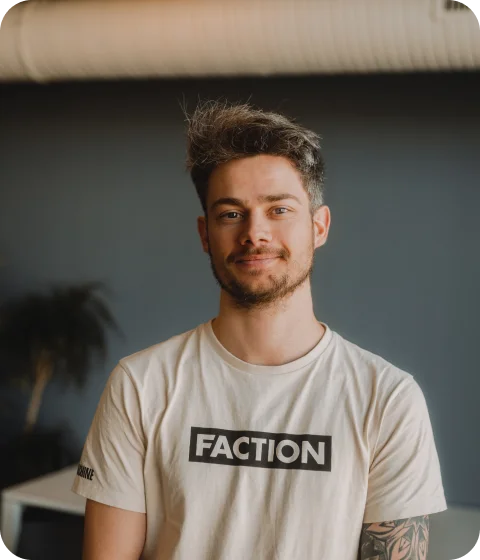 A man with short hair stands indoors, wearing a black sweatshirt and looking at the camera.