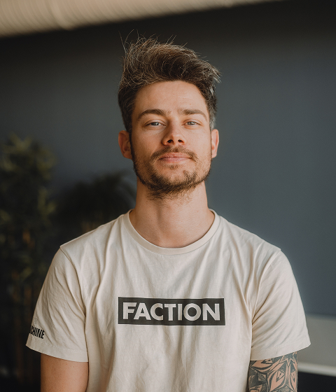 A man with short hair stands indoors, wearing a black sweatshirt and looking at the camera.
