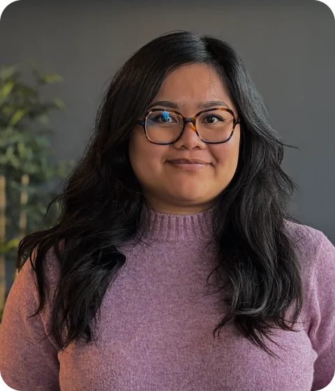 A woman with long dark hair and glasses is smiling, wearing a purple sweater.