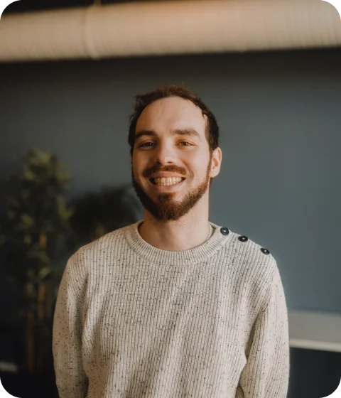 A person with glasses and a beard smiling, wearing a plaid shirt indoors.