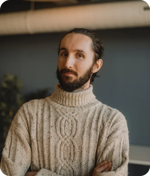 A person with a beard wearing a knitted sweater stands with arms crossed, looking at the camera.