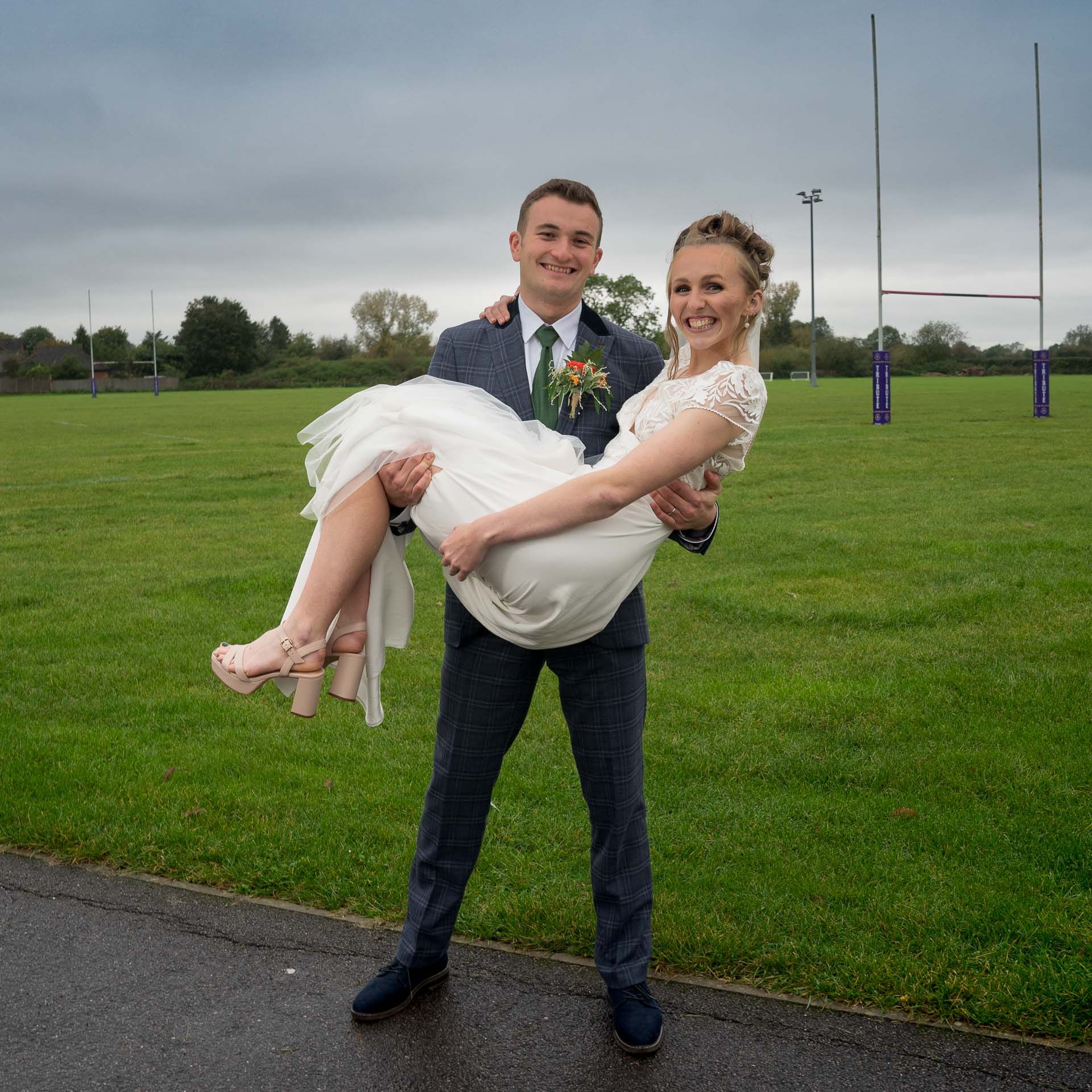 The groom easily carries the bride and stands in front of a rugby pitch