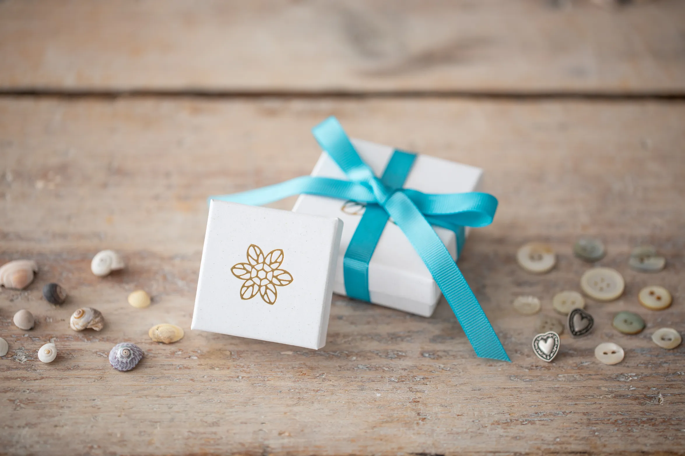 White gift box with a gold floral logo, tied with a blue ribbon, surrounded by small seashells and buttons on a wooden surface.