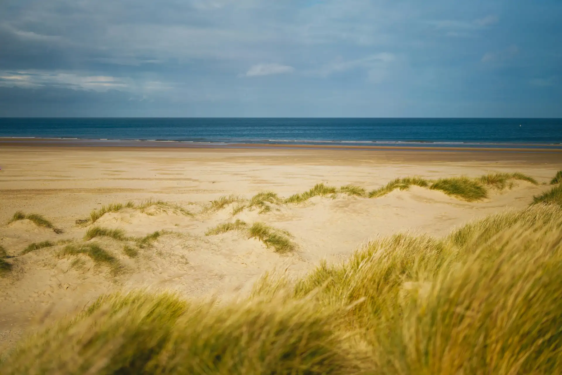 Sandy beach with grassy dunes in the foreground and calm ocean under a partly cloudy sky.