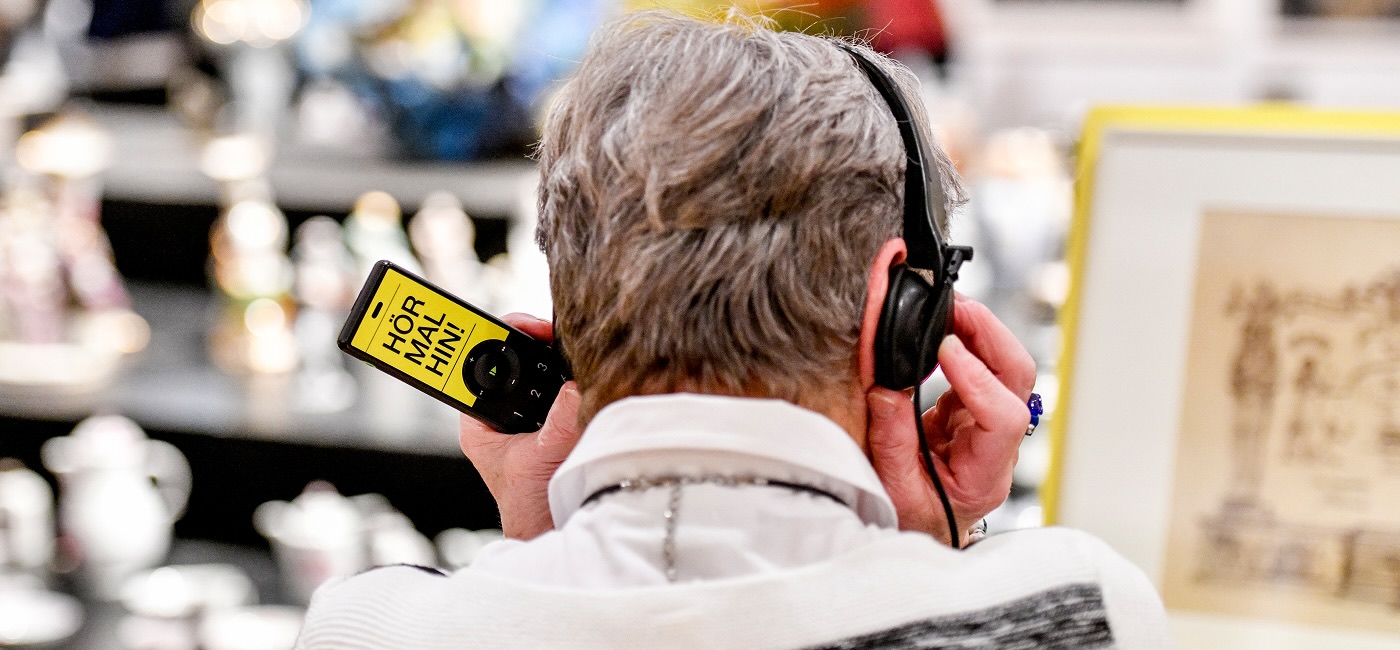Eine Person von hinten, in einem Museum, hält einen Audioguide mit der Aufschrift "HÖR MAL HIN!" und trägt Kopfhörer. Dies illustriert die Nutzung eines Trend Audioguides von Linon Medien in einer Ausstellung im Gustav-Lübcke Museum in Hamm. 