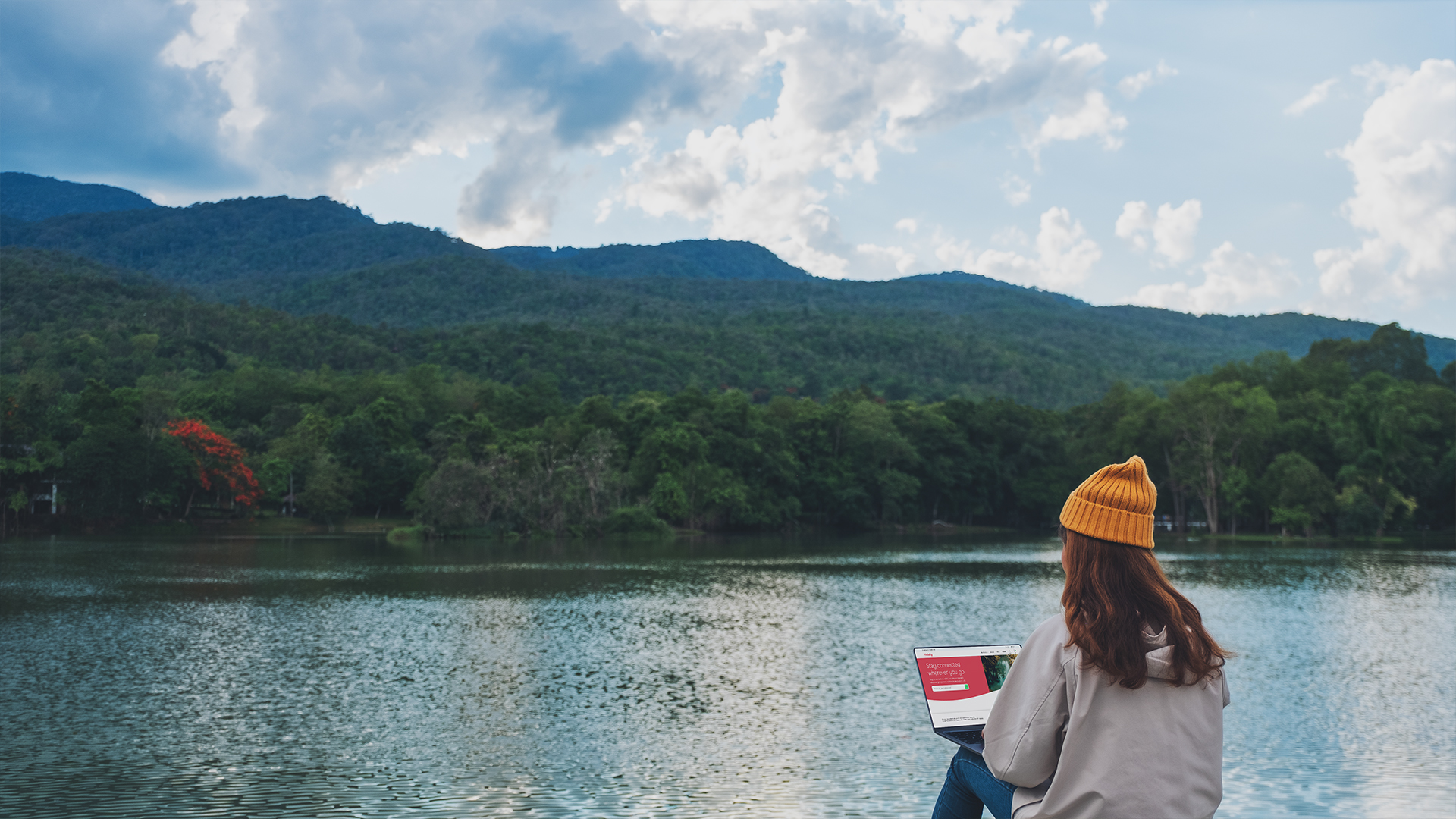 Woman in yellow beanie working on laptop by a mountain lake, with Holafly website visible on screen