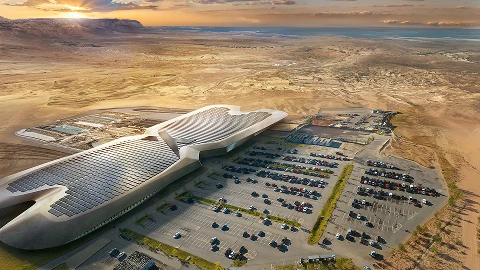 Aerial view of a large, modern building with solar panels on its roof, surrounded by a parking lot and arid landscape.