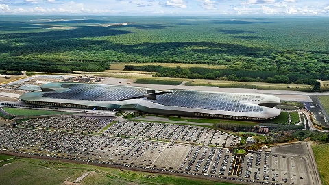 Aerial view of a large, modern building with solar panels on its roof, surrounded by a parking lot and arid landscape.
