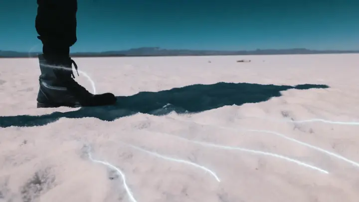 A person's boot stands on a white, expansive landscape with light streaks, casting a dark shadow under a clear sky.
