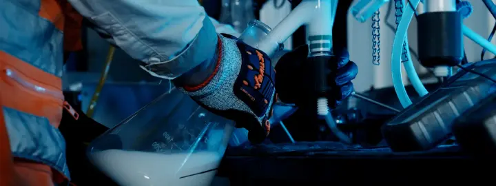 Person in gloves and lab coat pouring liquid from a large flask in a laboratory setting.