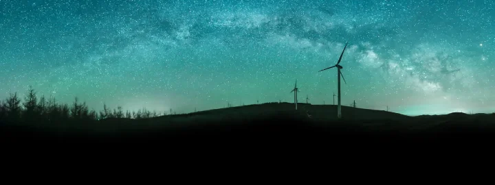 Silhouette of wind turbines on a hill under a starry, teal-colored night sky with the Milky Way visible.