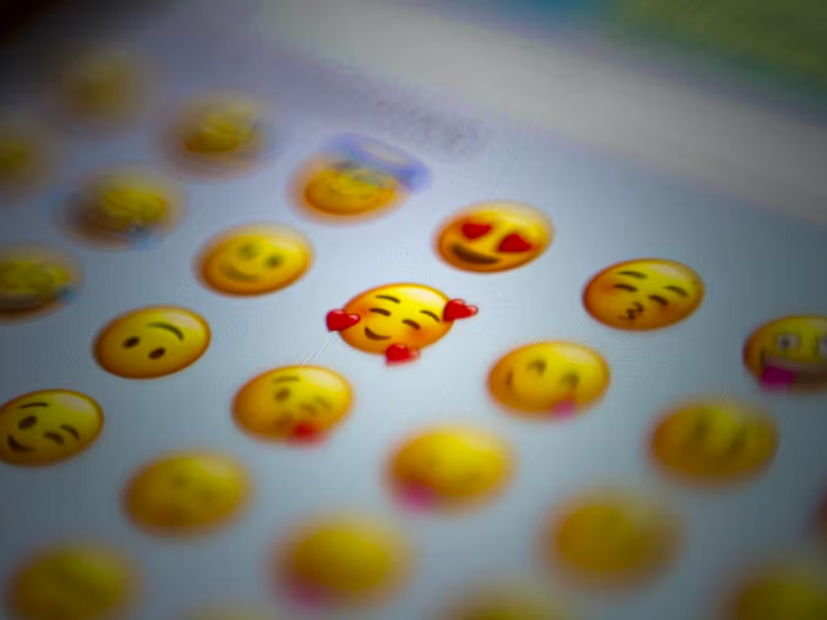 Blurred grid of emoji faces on a white screen, with a blushing smiley surrounded by hearts in focus, conveying a sense of love and affection.
