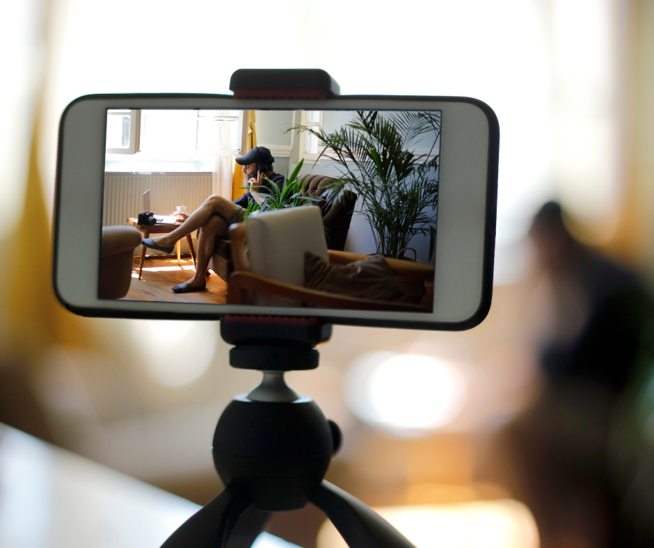 A smartphone on a tripod displays a person sitting on a sofa in a bright room, surrounded by plants. The scene feels calm and relaxed.