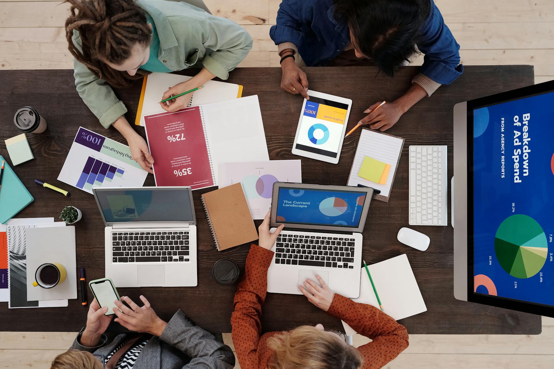 Aerial view of a diverse group collaborating at a table with laptops, tablets, charts, and coffee. The mood is focused and productive, emphasizing teamwork.