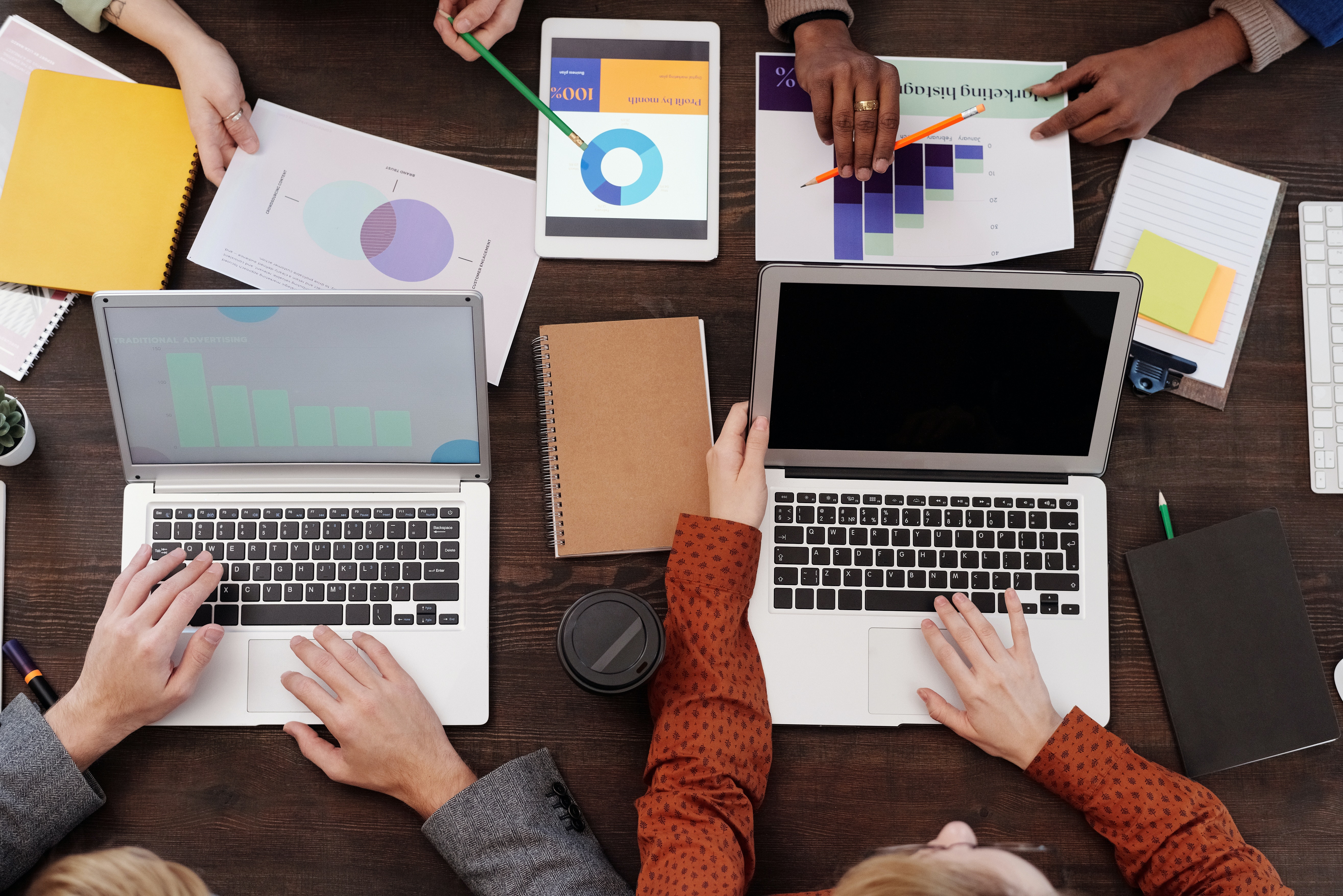 A diverse group collaborates around a table with laptops, tablets, and charts displaying graphs and data. The scene conveys teamwork and productivity.