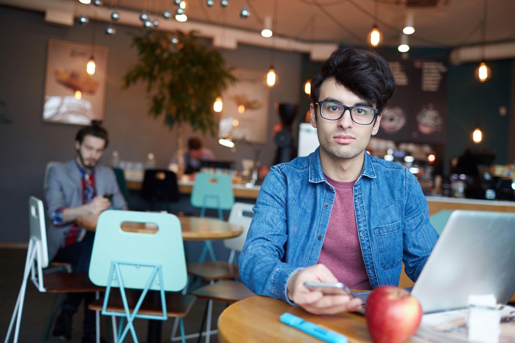 Young man in glasses sits at a café with a laptop and apple on the table, holding a phone. Cozy atmosphere with hanging lights and people in the background.