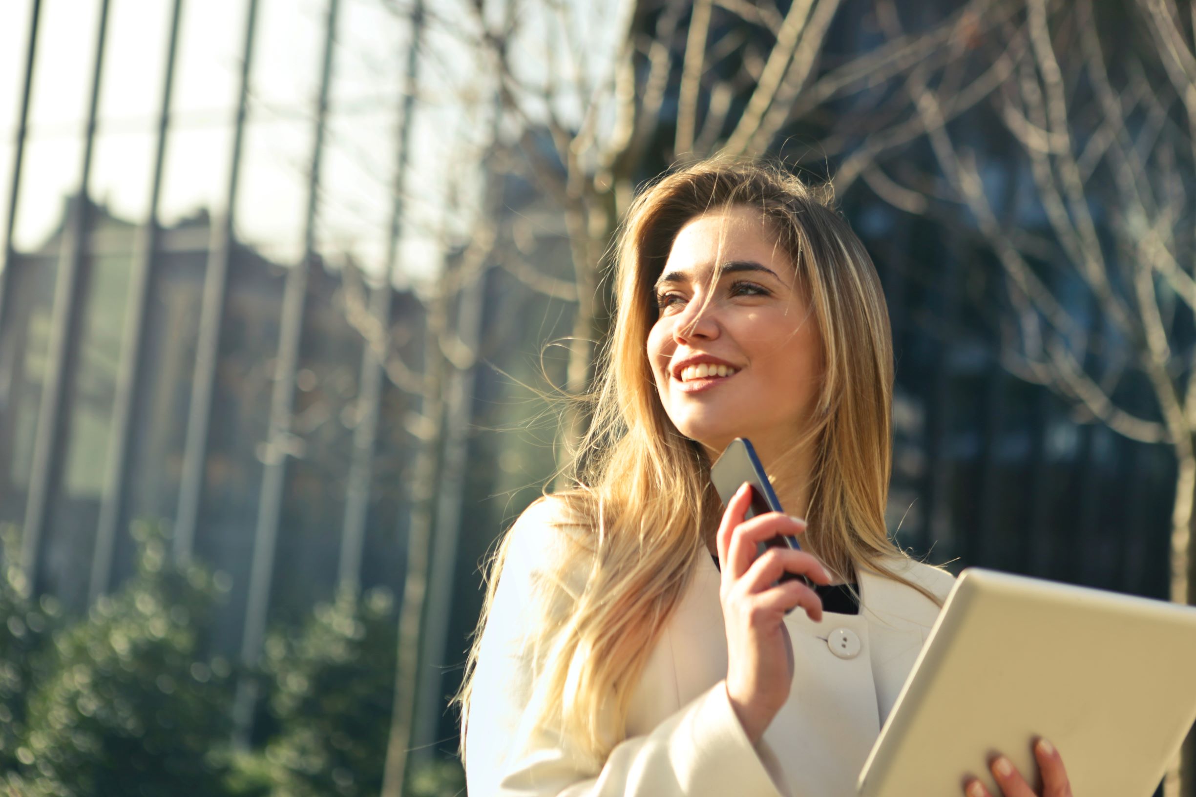 A woman smiles while holding a tablet and smartphone outdoors. She's dressed in white, standing in front of blurred trees and buildings, suggesting a sunny day.
