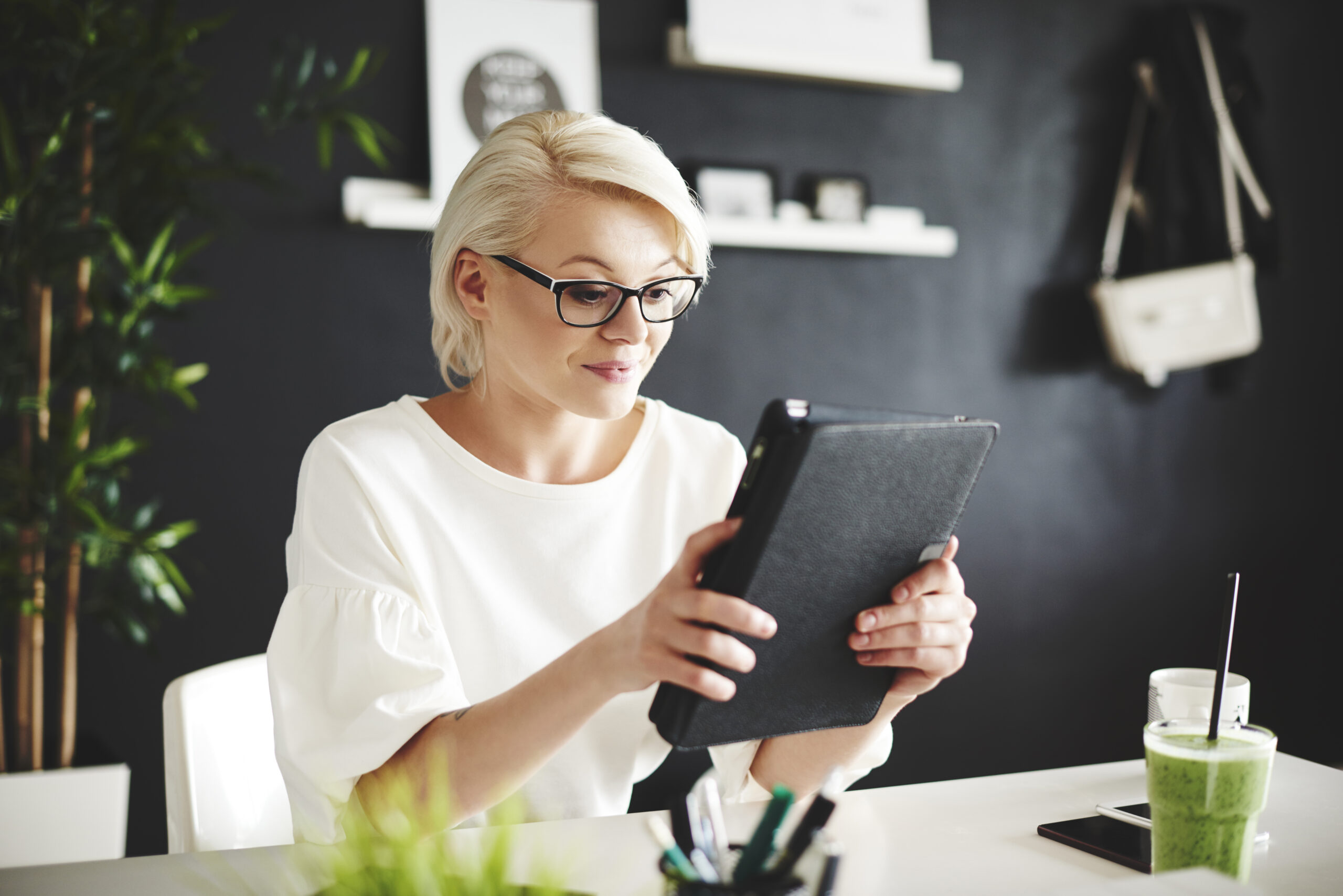 A woman with glasses uses a tablet at a modern desk with plants and shelves. She appears focused, and there's a green smoothie nearby.