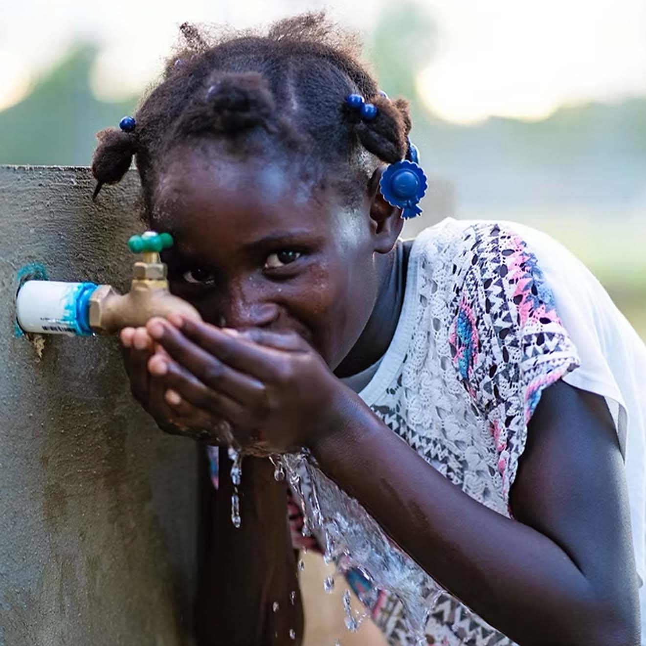 Young girl drinking water from an outdoor faucet using her hands.