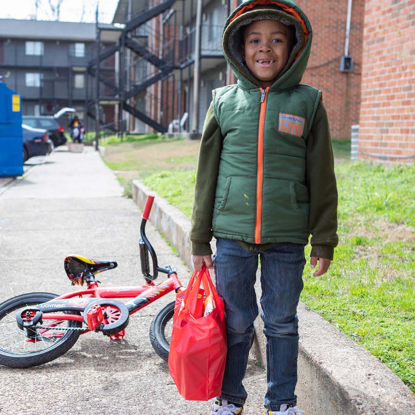 Young boy in a green hooded vest holding a red bag, standing on a sidewalk next to a red bicycle lying on its side.