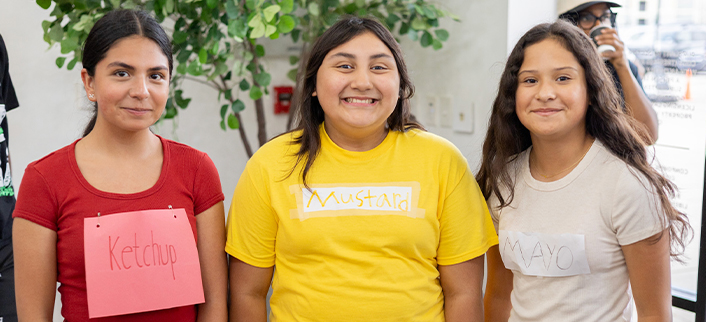 Three smiling girls wearing colored shirts with labels reading Ketchup, Mustard, and Mayo.