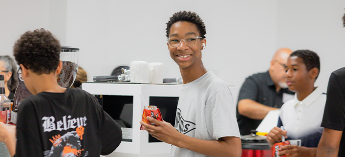 Teenage boy wearing glasses and a light gray shirt smiles while holding a snack pouch in a busy indoor setting with other youths.