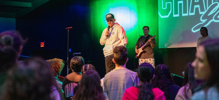 Man wearing a white hoodie and cap speaking into a microphone on stage with a guitarist and audience watching.