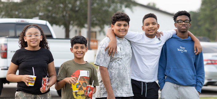 Five smiling students standing outdoors, some holding snacks and drinks, with two boys linking arms.
