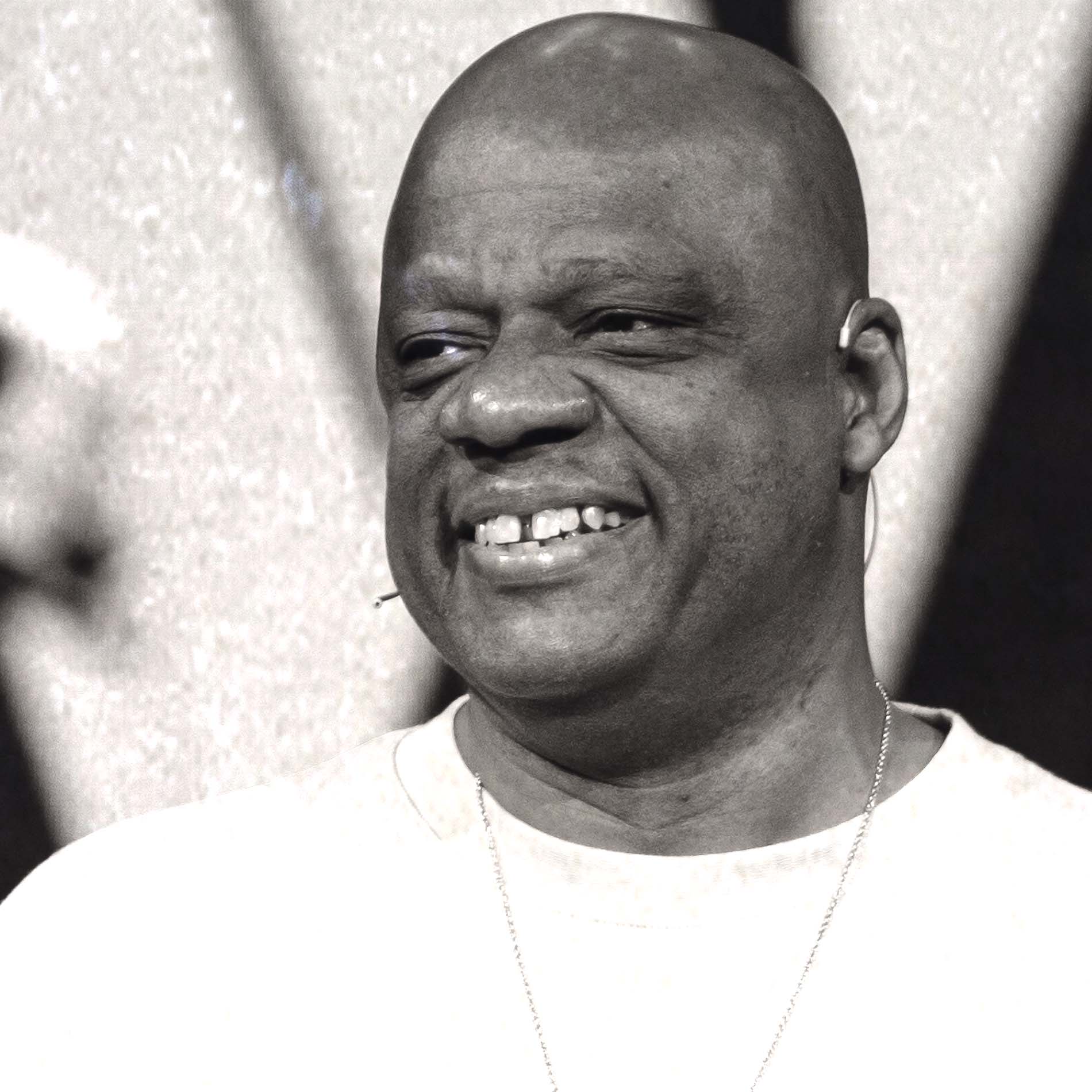 Smiling bald man with a headset mic, wearing a white shirt and chain necklace, looking to the side.