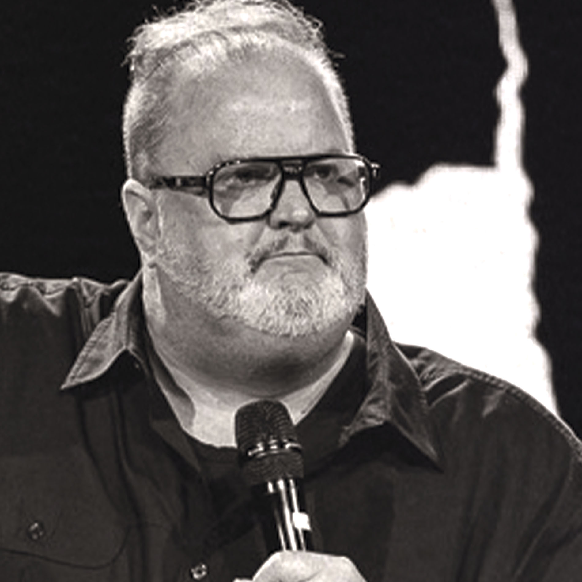 Man with glasses and white beard holding a microphone, wearing a black shirt, looking serious.