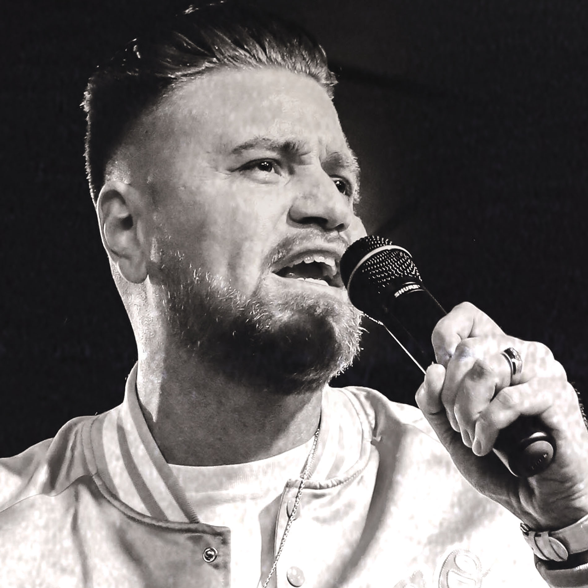 Bearded man passionately singing or speaking into a microphone on stage in black and white.