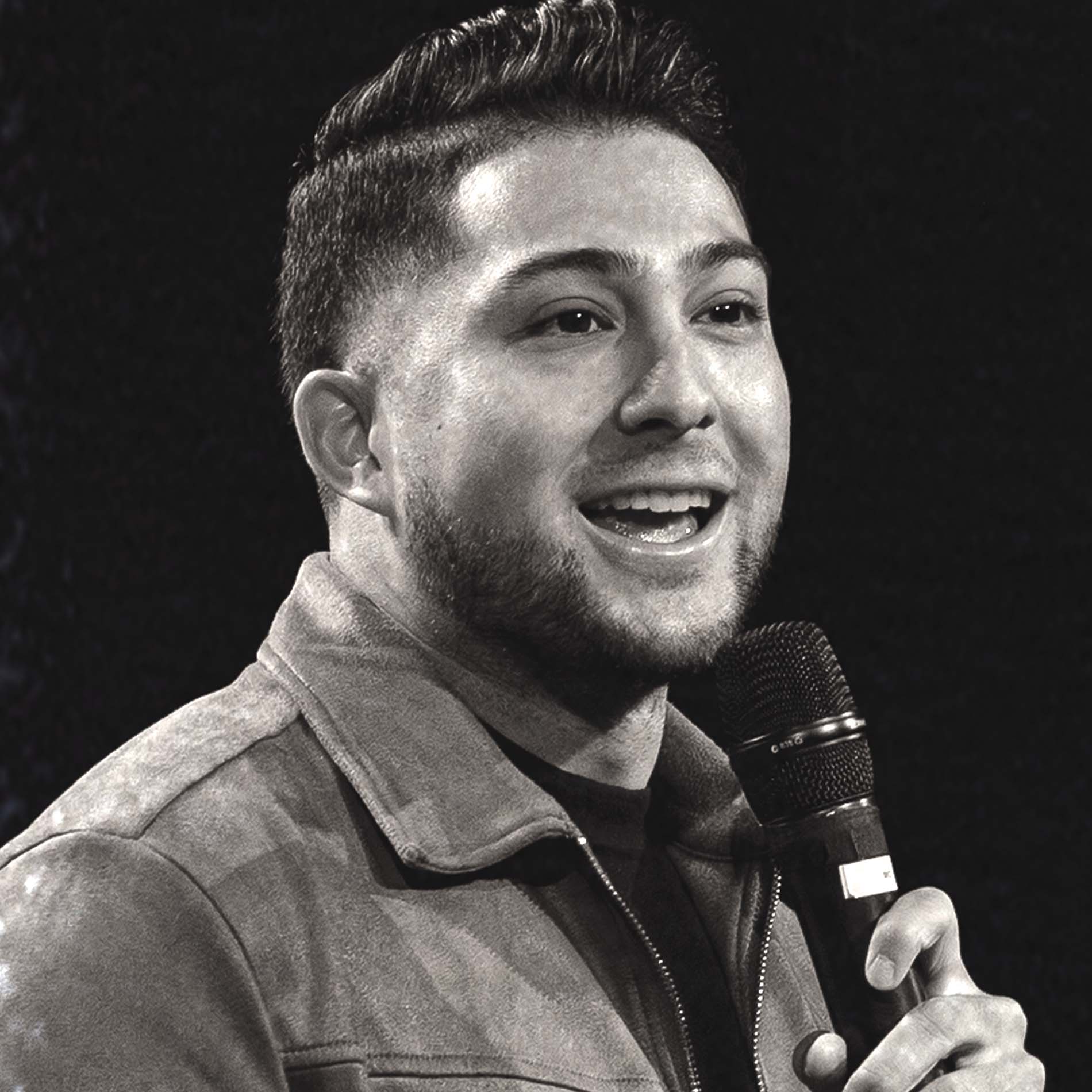 Smiling man speaking into a microphone with a dark background in black and white photo.