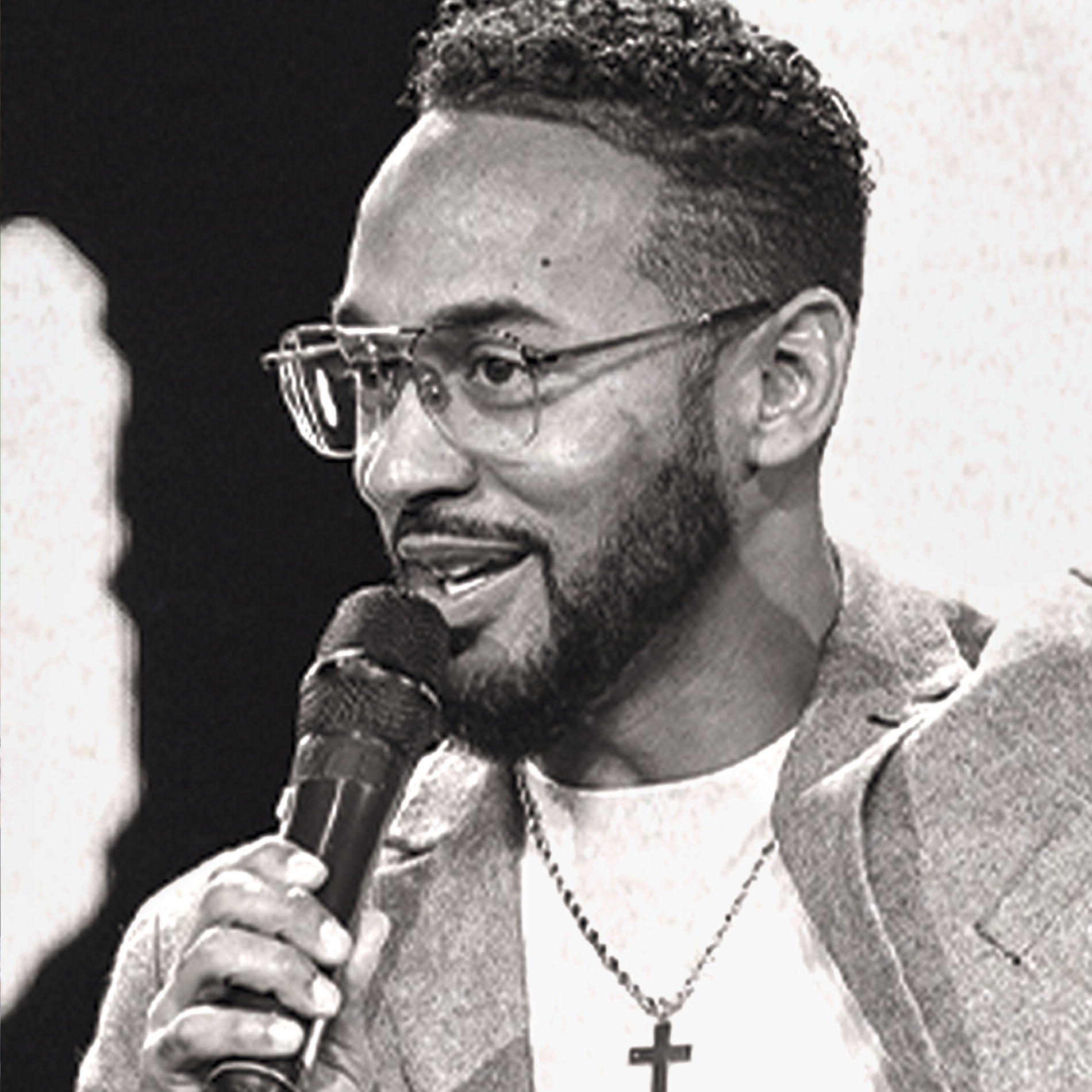 Man with glasses and beard speaks into microphone wearing cross necklace and blazer.