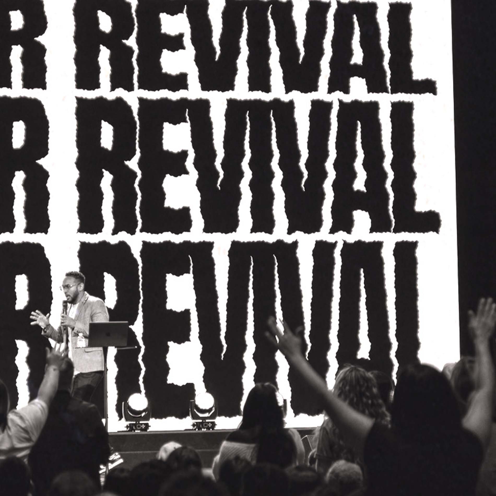 Speaker with microphone addresses an audience with raised hands in front of a large 'REVIVAL' sign.