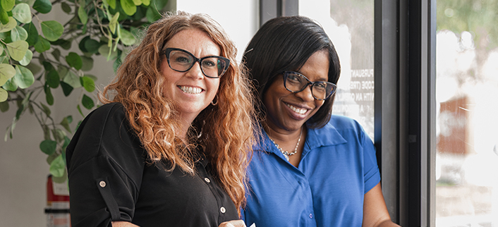 Two smiling women with glasses, one with curly red hair and the other with straight black hair.
