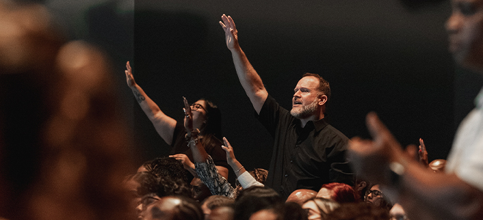People in a crowd raising hands, engaged and looking forward in a dark setting.