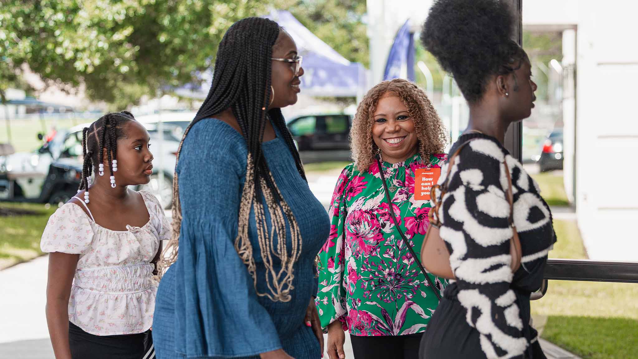 Four Black women standing outside, smiling and talking near a building entrance on a sunny day.