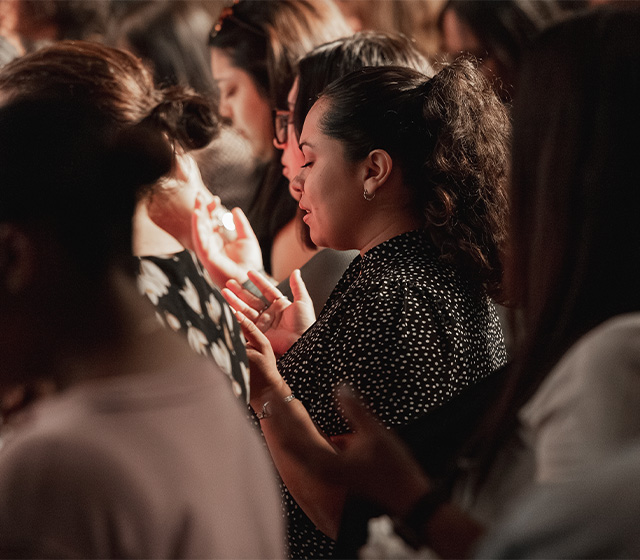 A group of people standing closely with eyes closed and hands raised, engaged in focused, collective prayer.