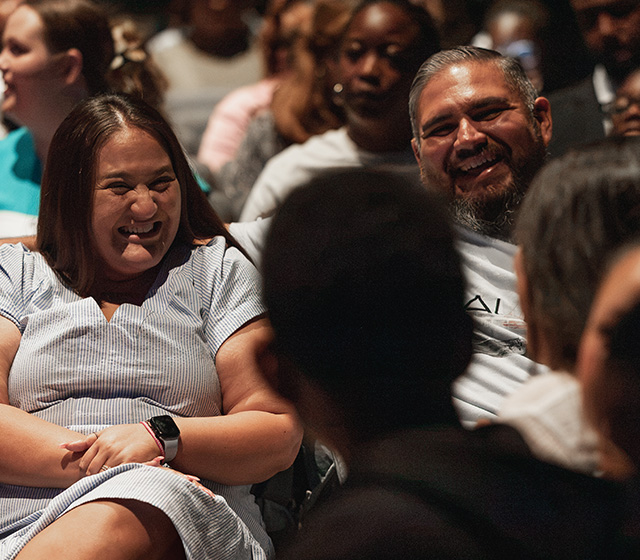 A group of people seated indoors, with two individuals in the foreground laughing and smiling while engaging with each other.